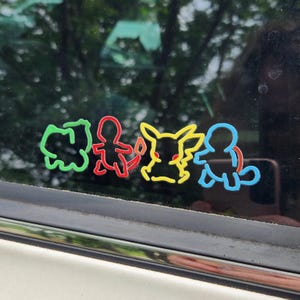 May include: A set of four colorful outline stickers featuring cartoon characters. The stickers are green, red, yellow, and blue, and are affixed to a window. The background shows a blurred view of trees.