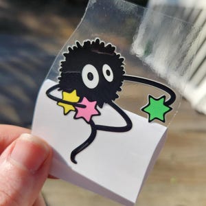 A close-up of a hand peeling a Ghibli-inspired sticker depicting a fuzzy black character with big eyes and thin limbs holding star-shaped objects, one yellow, one blue, and one pink.