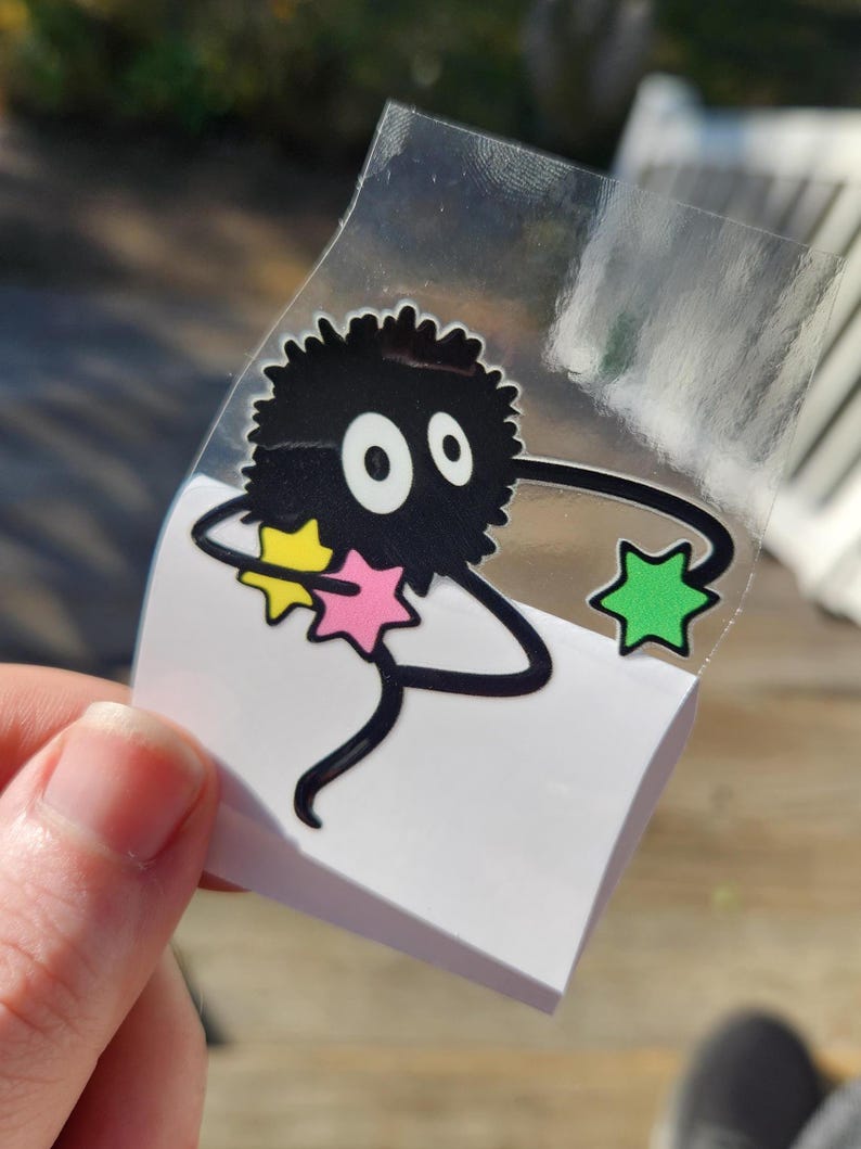 A close-up of a hand peeling a Ghibli-inspired sticker depicting a fuzzy black character with big eyes and thin limbs holding star-shaped objects, one yellow, one blue, and one pink.
