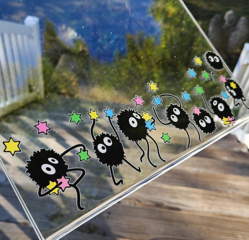 The glass panel showcases playful black cartoon creatures reminiscent of soot sprites, each clutching colorful stars. Stickers of these spirited figures dance across the surface, with a lush forest background visible through the transparent gaps.