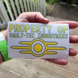 May include: A white sticker with the text "PROPERTY OF VAULT-TEC INDUSTRIES" in yellow and blue lettering. A yellow and blue Vault-Tec logo is below the text.