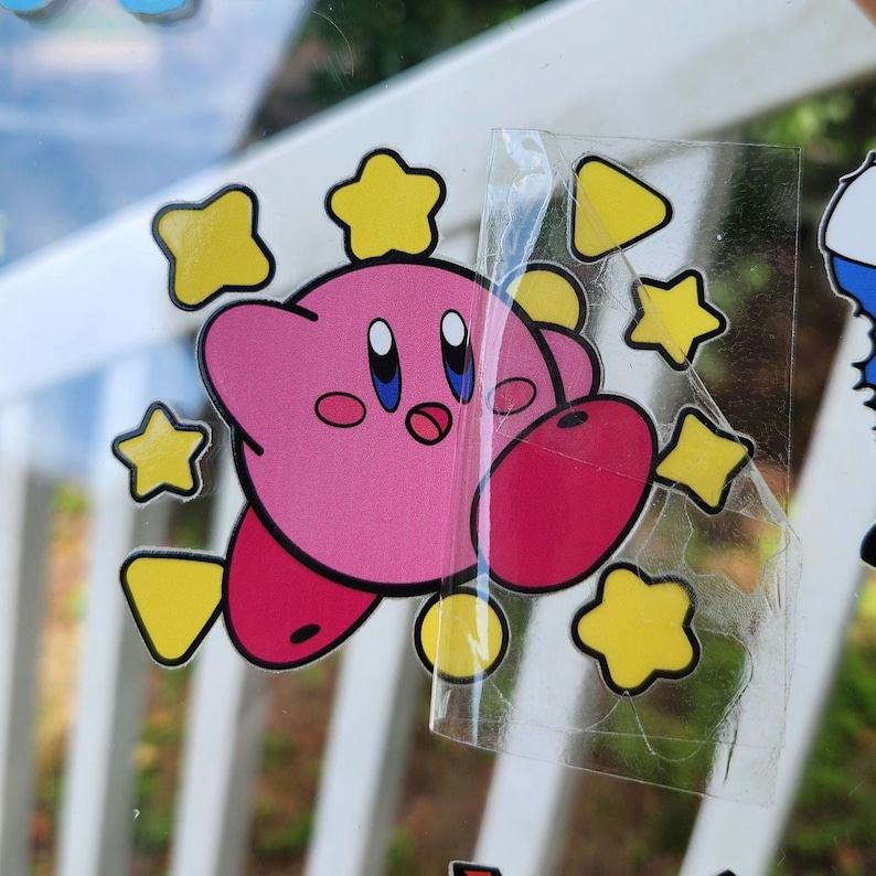 May include: Pink cartoon character with large eyes and a wide mouth, surrounded by yellow stars. The character is wearing a yellow star on its head. The image is a sticker.