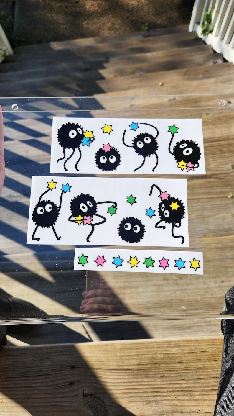The glass panel showcases playful black cartoon creatures reminiscent of soot sprites, each clutching colorful stars. A sticker sheet of these spirited figures dance across the surface, with a background visible through the transparent gaps.