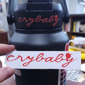 May include: Black water bottle with the word "crybaby" in red script on the bottle and a matching sticker. The sticker is held in front of the bottle. The bottle has a black handle.