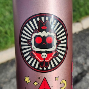 May include: A rose gold water bottle with a circular sticker featuring a cartoon lamb with red eyes and a red cloak. The sticker is surrounded by a black and white striped design. Additional stickers with stars and a red triangle are also visible.
