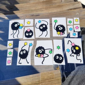 The glass panel showcases playful black cartoon creatures reminiscent of soot sprites, each clutching colorful stars. A sticker sheet of these spirited figures dance across the surface, with a background visible through the transparent gaps.