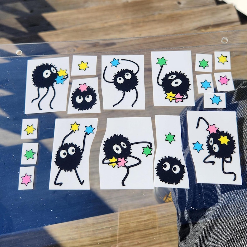 The glass panel showcases playful black cartoon creatures reminiscent of soot sprites, each clutching colorful stars. A sticker sheet of these spirited figures dance across the surface, with a background visible through the transparent gaps.