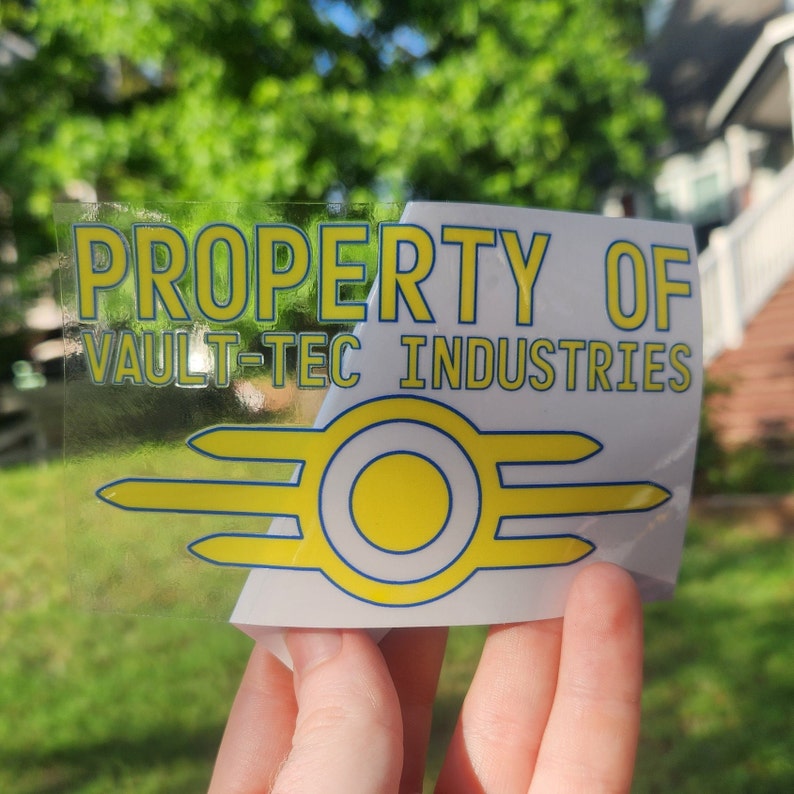 May include: A clear sticker with the text "PROPERTY OF VAULT-TEC INDUSTRIES" in yellow and blue lettering. A yellow and blue Vault-Tec logo is below the text.