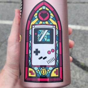 May include: A rose gold water bottle featuring a stained-glass style graphic of a retro handheld video game console. The design includes a heart, plus and minus buttons, and other game controls. The bottle is cylindrical and appears to be made of metal.