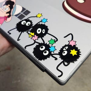 May include: A silver laptop with four black and white fuzzy round stickers with eyes and a string of colorful stars. The stickers are arranged in a line on the laptop.