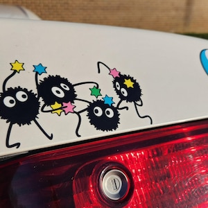 May include: A white car with a black and white sticker of a group of cartoon characters with large eyes. The characters are holding stars and are in a dancing pose. The car also has a sticker of a yin yang symbol.