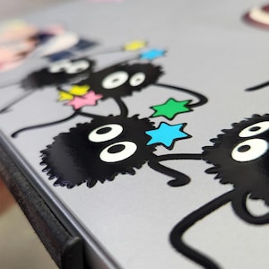 May include: A close-up of a gray laptop case with a black and white sticker of a line of fluffy black creatures with large eyes and stars. The creatures are holding hands and have a whimsical, cartoon-like appearance.
