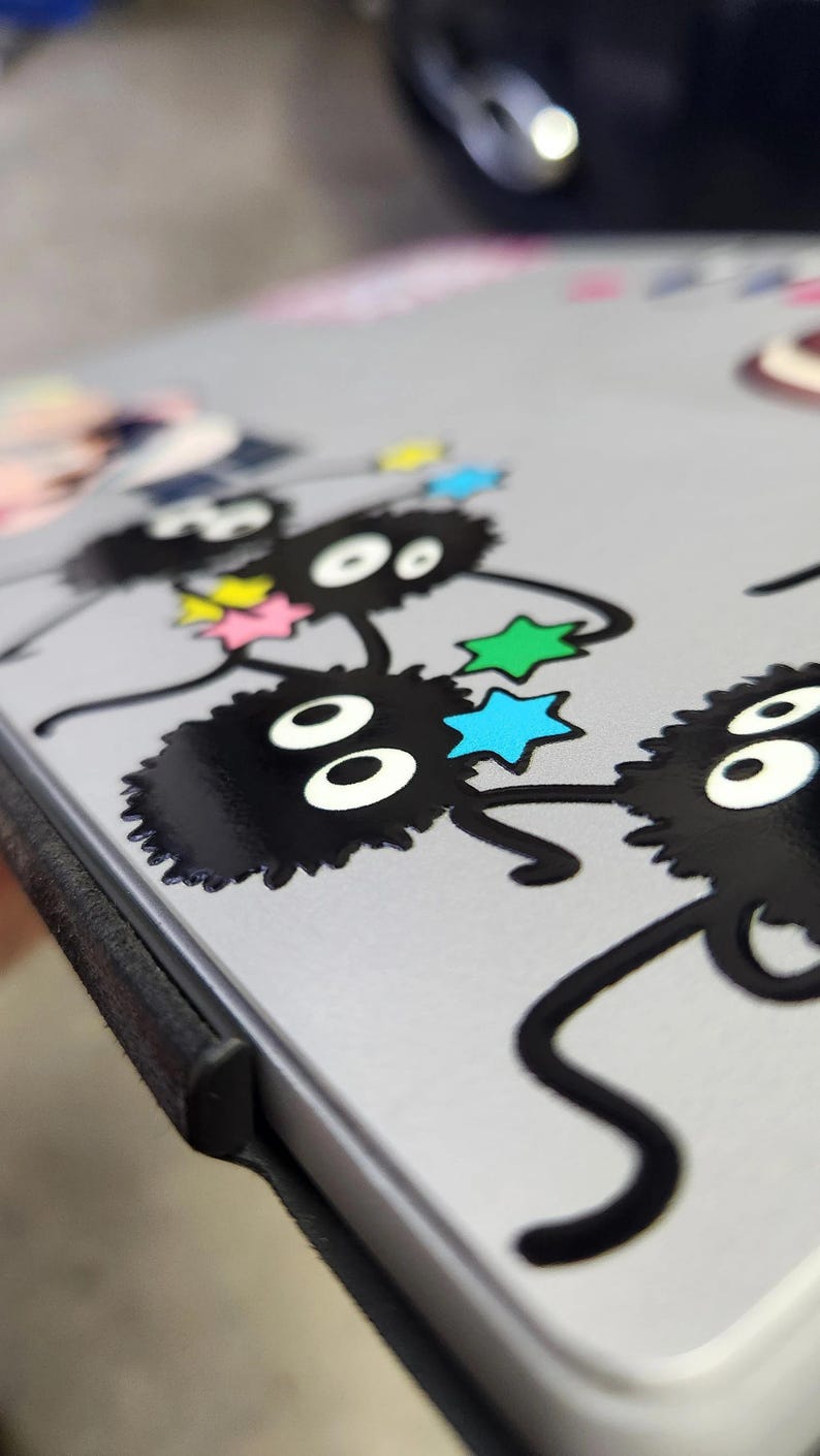 May include: A close-up of a gray laptop case with a black and white sticker of a line of fluffy black creatures with large eyes and stars. The creatures are holding hands and have a whimsical, cartoon-like appearance.