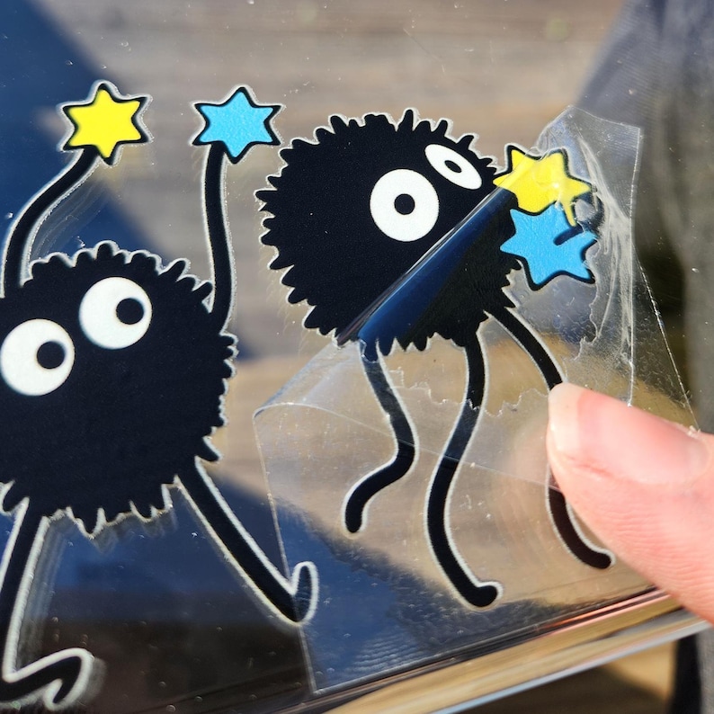 A close-up of a hand peeling a Ghibli-inspired sticker depicting a fuzzy black character with big eyes and thin limbs holding star-shaped objects, one yellow and one blue.