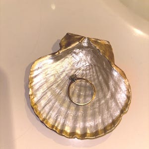 May include: A gold and silver seashell-shaped trinket dish with a gold ring. The dish has a metallic finish with gold edging. The ring has a gold band and a clear gemstone.