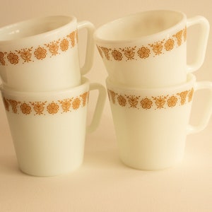 Four matching white glass mugs stacked with diagonal shaped handles and orange-gold butterfly details around each rim