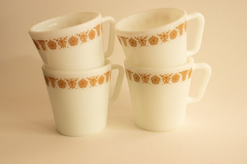 Four matching white glass mugs stacked with diagonal shaped handles and orange-gold butterfly details around each rim