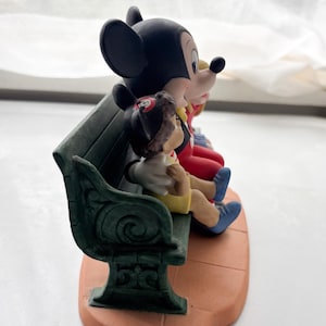 K&ouml;nnte beinhalten: Eine Keramikfigur mit Mickey Mouse, Minnie Mouse und Pinocchio auf einer gr&uuml;nen Bank. Mickey tr&auml;gt rote Shorts und Schuhe, Minnie hat eine rote Schleife und Pinocchio tr&auml;gt gelbe Shorts. Der Sockel ist terrakottafarben.