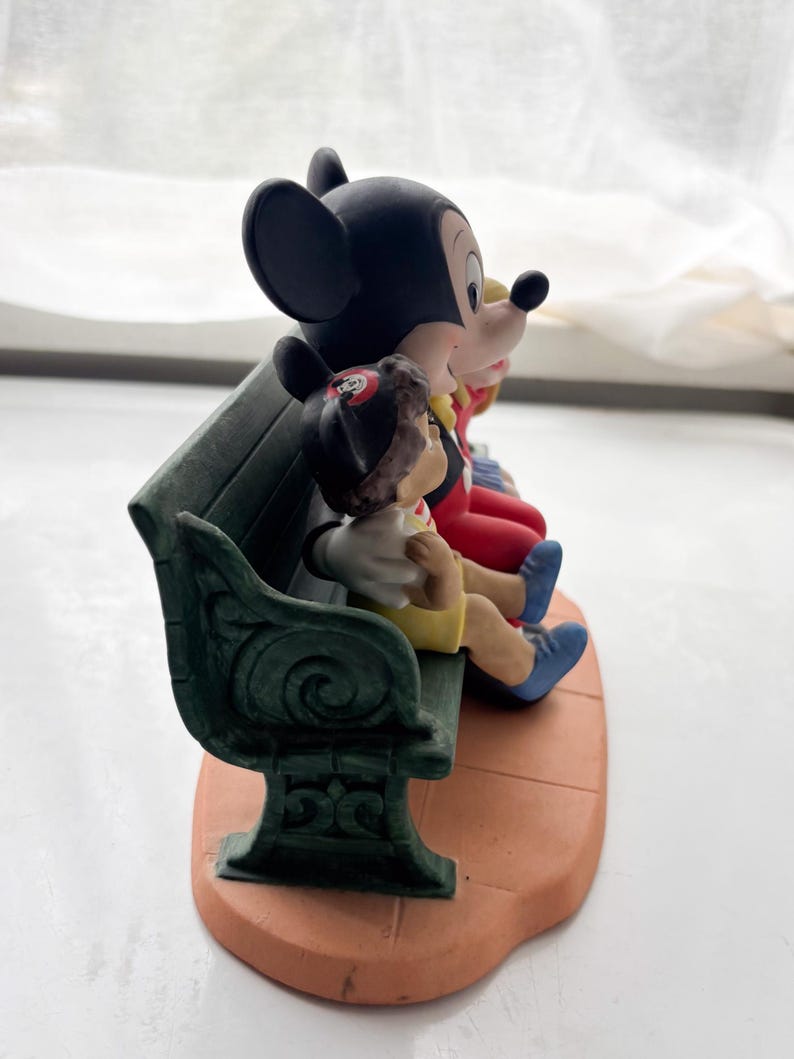K&ouml;nnte beinhalten: Eine Keramikfigur mit Mickey Mouse, Minnie Mouse und Pinocchio auf einer gr&uuml;nen Bank. Mickey tr&auml;gt rote Shorts und Schuhe, Minnie hat eine rote Schleife und Pinocchio tr&auml;gt gelbe Shorts. Der Sockel ist terrakottafarben.