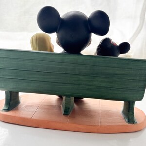 K&ouml;nnte beinhalten: Eine Figur mit Mickey Mouse, Minnie Mouse und einer blonden Figur, die auf einer gr&uuml;nen Bank sitzen. Die Bank steht auf einem orangefarbenen Sockel. Die Figuren sind von hinten zu sehen.