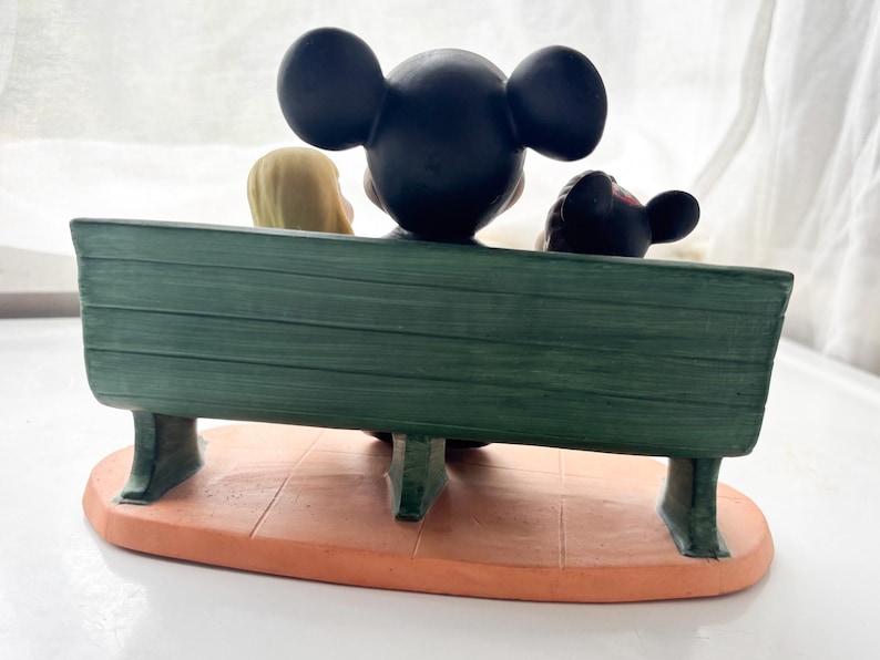 K&ouml;nnte beinhalten: Eine Figur mit Mickey Mouse, Minnie Mouse und einer blonden Figur, die auf einer gr&uuml;nen Bank sitzen. Die Bank steht auf einem orangefarbenen Sockel. Die Figuren sind von hinten zu sehen.