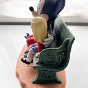 K&ouml;nnte beinhalten: Eine Keramikfigur von Mickey und Minnie Mouse auf einer gr&uuml;nen Bank. Mickey, in einem schwarzen Anzug, umarmt Minnie, die blondes Haar und ein rotes Kleid hat. Der Sockel ist terrakottafarben, und die Bank hat dekorative Details.