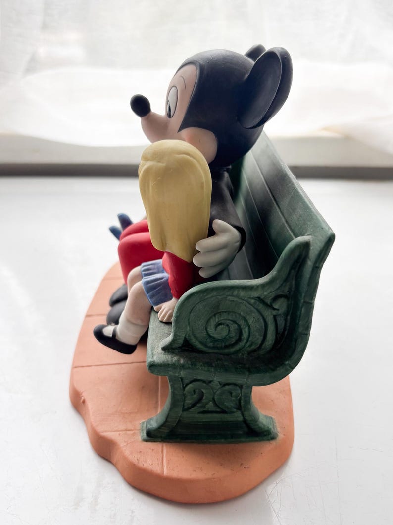 K&ouml;nnte beinhalten: Eine Keramikfigur von Mickey und Minnie Mouse auf einer gr&uuml;nen Bank. Mickey, in einem schwarzen Anzug, umarmt Minnie, die blondes Haar und ein rotes Kleid hat. Der Sockel ist terrakottafarben, und die Bank hat dekorative Details.