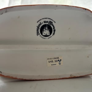 K&ouml;nnte beinhalten: Wei&szlig;er Keramik-Disney-Figurensockel mit braunem Rand. Der Sockel hat ein Disneyland- und Walt Disney World-Logo. Der Text "DESIGNED AND MADE ESPECIALLY FOR" ist &uuml;ber dem Logo aufgedruckt.