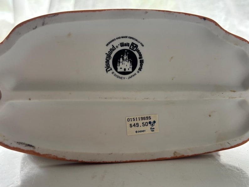 K&ouml;nnte beinhalten: Wei&szlig;er Keramik-Disney-Figurensockel mit braunem Rand. Der Sockel hat ein Disneyland- und Walt Disney World-Logo. Der Text "DESIGNED AND MADE ESPECIALLY FOR" ist &uuml;ber dem Logo aufgedruckt.