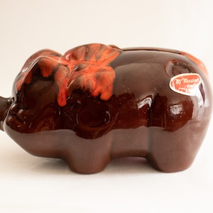 May include: A ceramic piggy bank shaped like a pig, with a glossy brown finish and red detailing. The piggy bank has a slot for coins and a sticker that reads "M' Master craft".