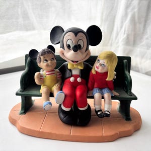 K&ouml;nnte beinhalten: Keramikfigur mit Mickey Mouse auf einer gr&uuml;nen Bank mit zwei Kindern. Mickey tr&auml;gt eine schwarze Jacke, eine rote Hose und eine gelbe Fliege. Die Kinder tragen gelbe und rote Kleidung. Der Sockel ist hellbraun.