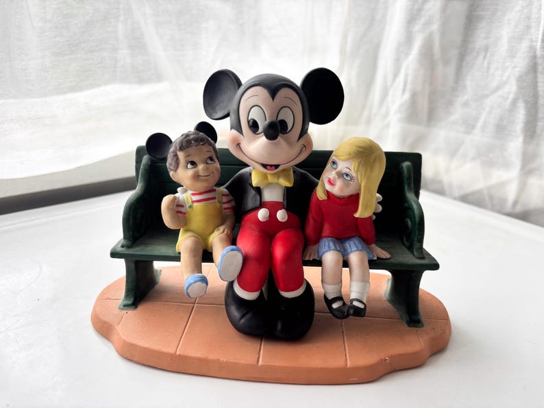 K&ouml;nnte beinhalten: Keramikfigur mit Mickey Mouse auf einer gr&uuml;nen Bank mit zwei Kindern. Mickey tr&auml;gt eine schwarze Jacke, eine rote Hose und eine gelbe Fliege. Die Kinder tragen gelbe und rote Kleidung. Der Sockel ist hellbraun.