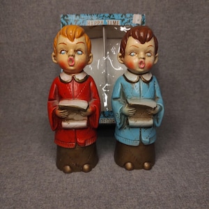 Puede incluir: Dos figuras de niños de coro de estilo vintage frente a su caja original con la etiqueta Happy Time. Una figura lleva una chaqueta roja y la otra una azul, ambas con cuellos blancos, faldas marrones y sosteniendo libros de canciones abiertos.