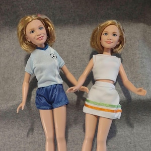 Puede incluir: Dos muñecas de cabello rubio. La muñeca de la izquierda viste una camiseta azul claro de manga corta con un gráfico de balón de fútbol y pantalones cortos azules. La muñeca de la derecha viste un top corto blanco sin mangas y una falda blanca con ribetes verdes y naranjas.