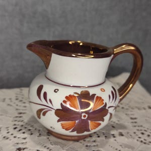 May include: Small white ceramic creamer with a brown and orange floral design. The creamer has a gold-colored handle and rim. The floral design is centered on the front of the creamer. The creamer is sitting on a white lace tablecloth.