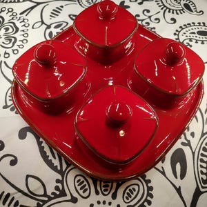 Könnte beinhalten: Ein rotes Keramik-Servierset mit vier quadratischen Schalen mit Deckel auf einem passenden Tablett. Jede Schale hat einen kleinen runden Knopf auf dem Deckel. Das Set hat eine glänzende Oberfläche und einen braunen Rand.