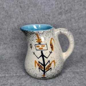 May include: Small ceramic jug with a white speckled finish and a blue interior. The jug features a black and orange abstract design of a figure with a square head and a handle.