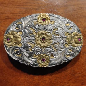 May include: An oval belt buckle featuring a two-tone silver and gold finish with intricate floral scrollwork engravings. The design includes five small, circular, deep red stones embedded in the centers of the gold-toned flower motifs.