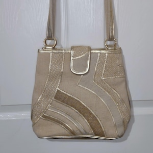 Könnte beinhalten: Eine beigefarbene Umhängetasche mit goldenen Akzenten. Die Tasche hat einen Klappverschluss und einen langen Riemen. Goldene Verzierungen und dekorative Muster zieren die Vorderseite der Tasche. Die Tasche ist aus einem strukturierten Stoff gefertigt.