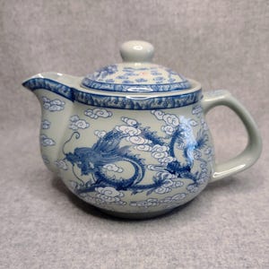 May include: A light grey ceramic teapot featuring a blue painted dragon motif surrounded by stylised clouds. The teapot includes a matching lid with a knob handle, a curved spout, and a side handle, all decorated with blue cloud patterns.