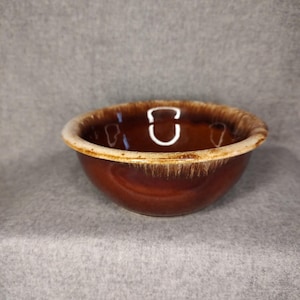 May include: A brown ceramic bowl with a cream-colored rim. The bowl has a glossy finish and a rounded shape. The interior is a darker shade of brown. This bowl is suitable for serving food.
