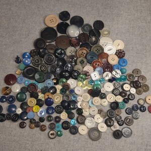 May include: A collection of various buttons in different sizes, shapes, and colors. The buttons include shades of black, brown, white, blue, and green. Some buttons have multiple holes, while others have a shank.