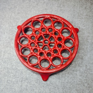 May include: A circular red trivet featuring an intricate openwork pattern of interconnected rings and circles of varying sizes. The design includes a central hexagonal arrangement surrounded by multiple concentric rings, supported by three small feet.