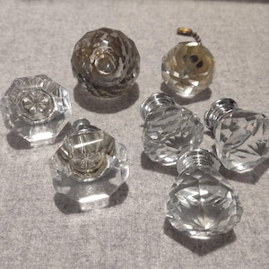 May include: A collection of six clear, multifaceted glass cabinet knobs with a geometric, diamond-like shape. Each knob features a polished silver-tone metal base and a reflective surface that catches light, creating a sparkling, transparent appearance.