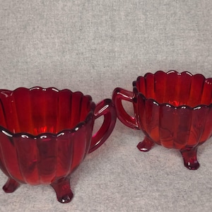 May include: A ruby red glass creamer and sugar bowl set. The creamer has a spout and a handle, while the sugar bowl has two handles. Both pieces have a scalloped rim and sit on three small feet. The set is made of a translucent red glass.