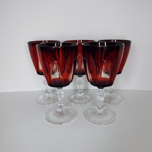 May include: A set of five wine glasses featuring deep red bowls with vertical panel cuts and clear glass stems and circular bases. The glasses are arranged in two rows, with three in the back and two in the front, showcasing the dark ruby color.