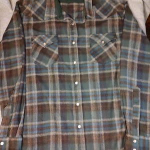 May include: A long-sleeved plaid shirt in shades of blue, brown, and green. The shirt features a green collar and two chest pockets with button closures. The shirt has white buttons down the front and on the cuffs. The brand name "Pendleton" is visible on the collar.