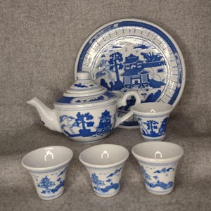 Peut inclure: Service à thé en porcelaine blanche et bleue comprenant une théière, une assiette et quatre petites tasses. L'ensemble présente un design asiatique traditionnel avec des illustrations de paysages bleus. La théière a un bec courbé et un couvercle arrondi.