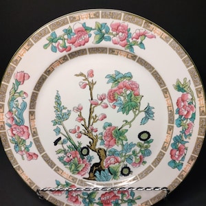 May include: A white ceramic plate with a floral design. The plate features pink flowers, green leaves, and blue accents. A gold border with a geometric pattern encircles the plate's edge. The plate is displayed on a silver metal stand.