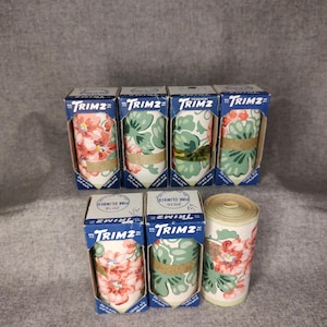 Pode incluir: Vários rolos de borda de papel de parede TRIMZ vintage em caixas. As caixas são azuis e brancas com o logotipo TRIMZ. O papel de parede apresenta padrões florais em rosa, verde e branco. Alguns rolos são amarrados com barbante.